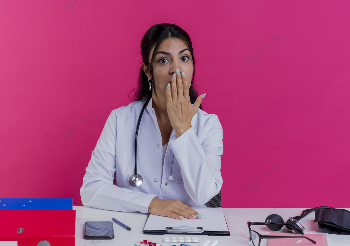 工具惊讶的年轻女医生穿着医用长袍和听诊器坐在办公桌旁 手放在办公桌上 嘴被隔离在粉红色的墙上坐着医生长袍