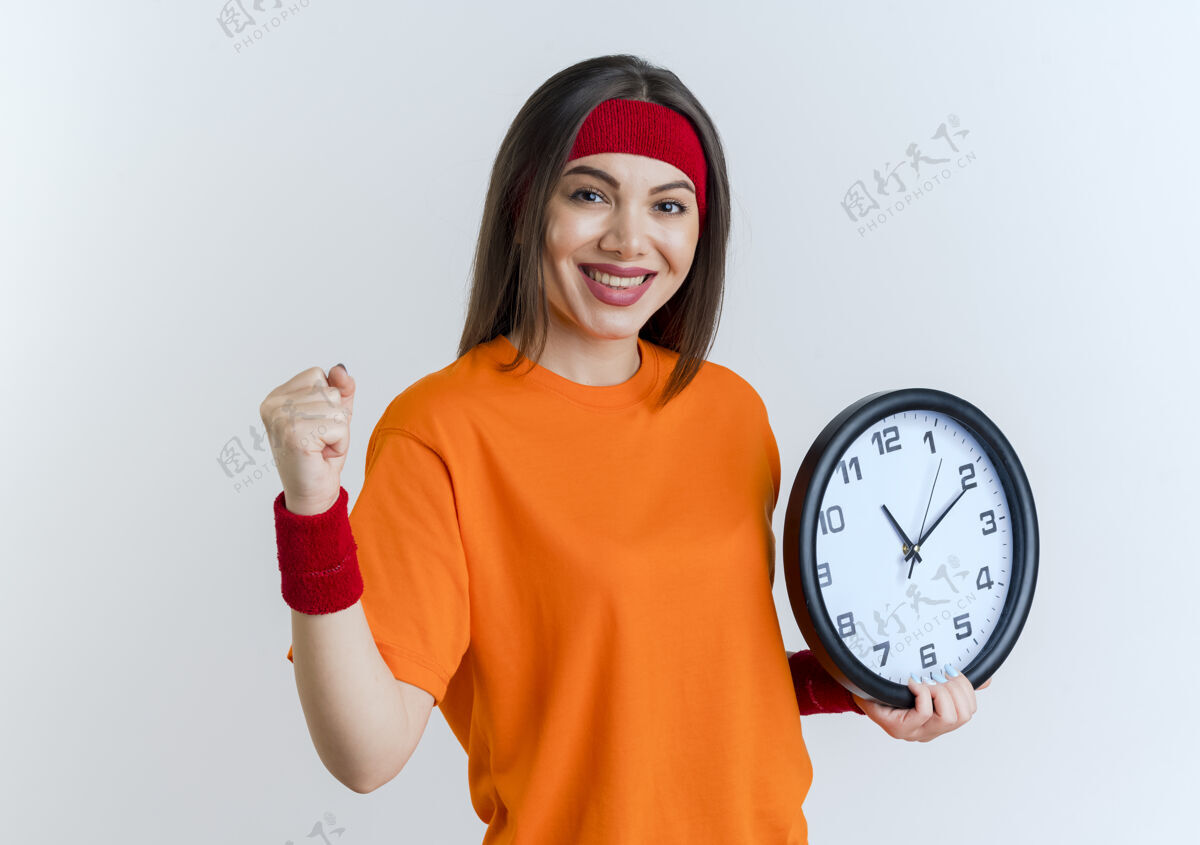 时钟微笑的年轻运动女性戴着头带和腕带拿着时钟做着被隔离在白墙上的强烈手势握着手势腕带