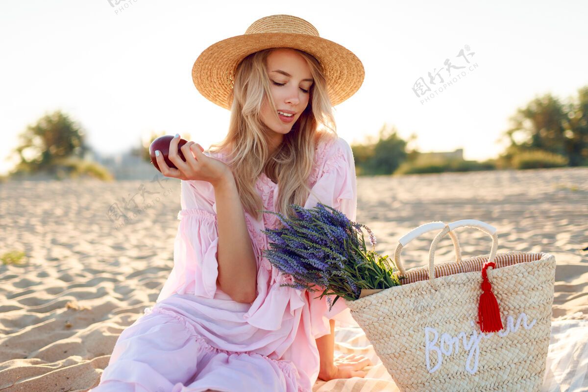 食物在海边的乡间野餐优雅的年轻女子 金色卷发 穿着优雅的粉色连衣裙 享受假期 吃水果金发女孩坐着