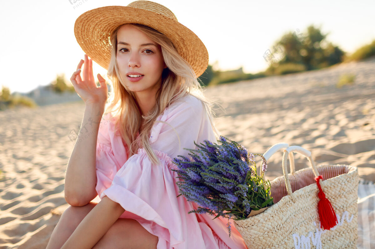 野餐浪漫的白人女子 戴着时髦的帽子 穿着优雅的粉色连衣裙 手持草包和花束 在沙滩上摆着姿势肖像苹果健康