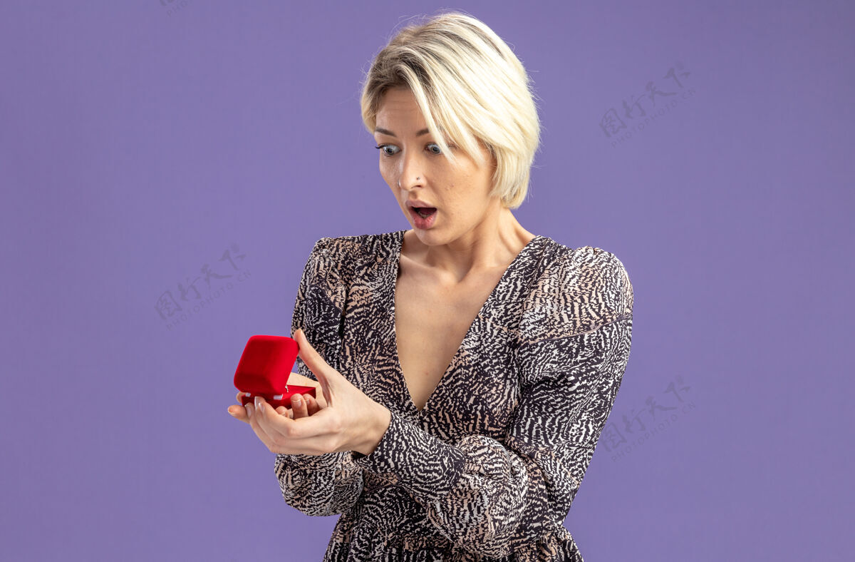立场年轻漂亮的女人穿着礼服拿着红色的订婚戒指看着它惊讶和惊喜的情人节概念站在紫色的墙上订婚惊喜日子