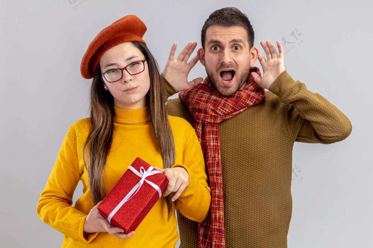 脸年轻漂亮的夫妇兴奋的男子和微笑的女子在贝雷帽举行的礼物 脸上带着微笑庆祝情人节站在白色的墙壁贝雷帽兴奋情人节