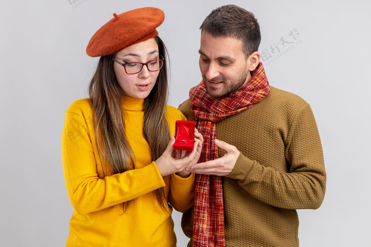 年轻年轻漂亮的夫妇幸福的男人和微笑的女人在贝雷帽与订婚戒指在红盒子庆祝情人节站在白色的背景戒指情侣订婚