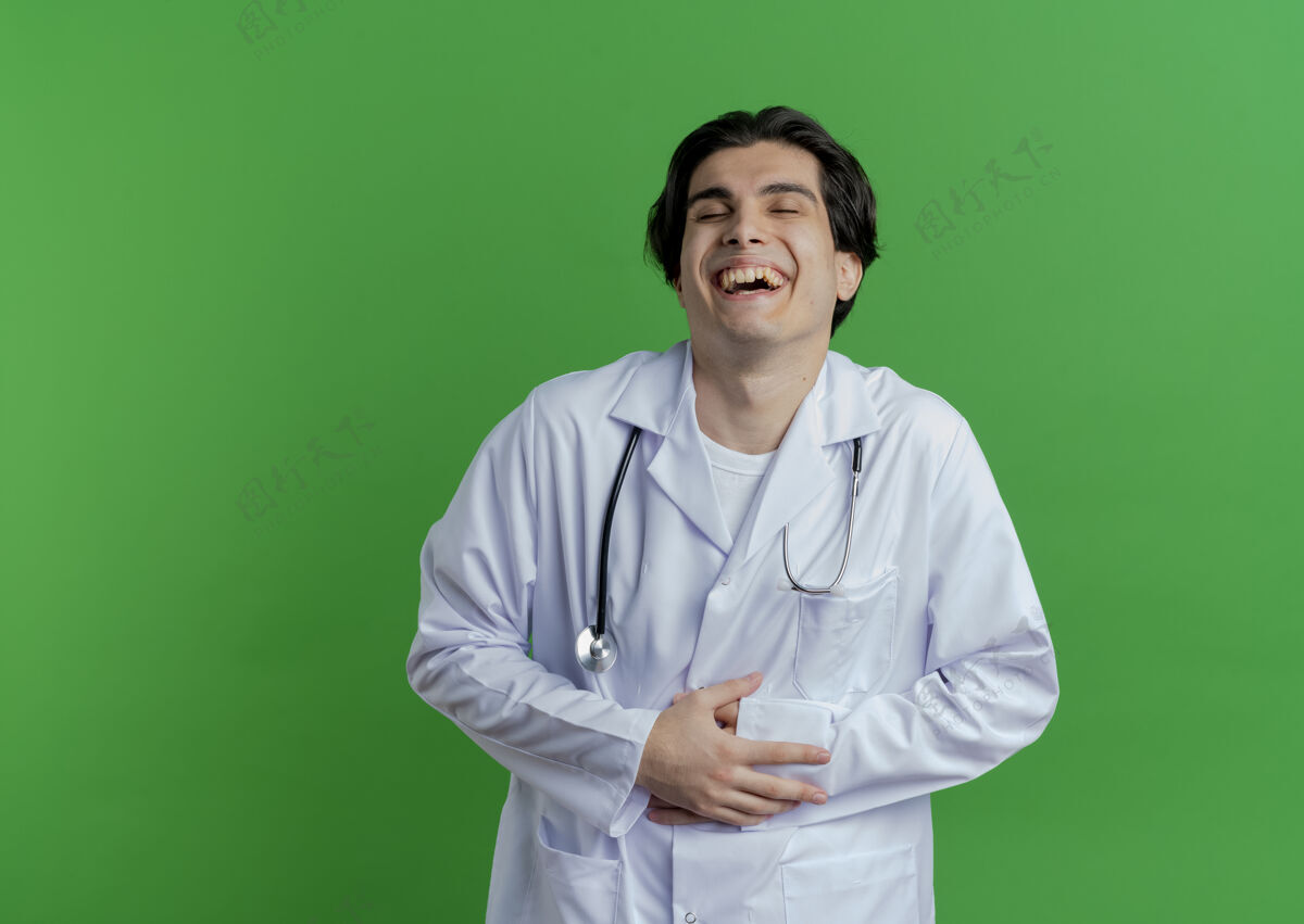 抱着年轻的男医生穿着医用长袍和听诊器抱着肚子笑着闭着眼睛隔离在有复印空间的墙上男性肚子年轻