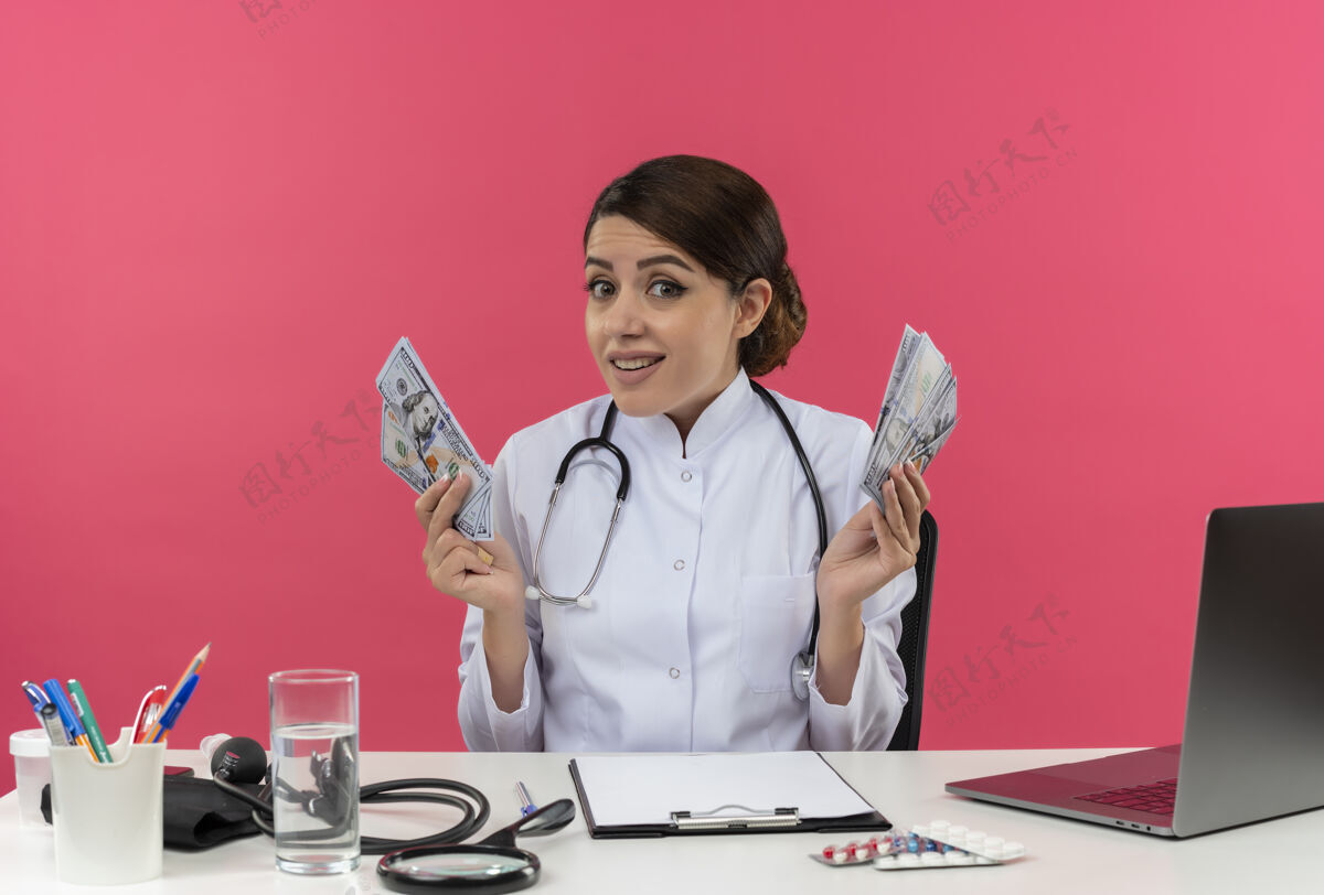 坐着年轻的女医生穿着医用长袍和听诊器坐在办公桌旁 手里拿着医疗工具和笔记本电脑 把钱隔离在粉红色的墙上穿着办公桌印象