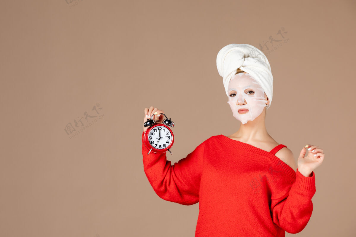 润肤霜正面图：戴着面具的年轻女性 手持粉色背景上的时钟帽子面具脸