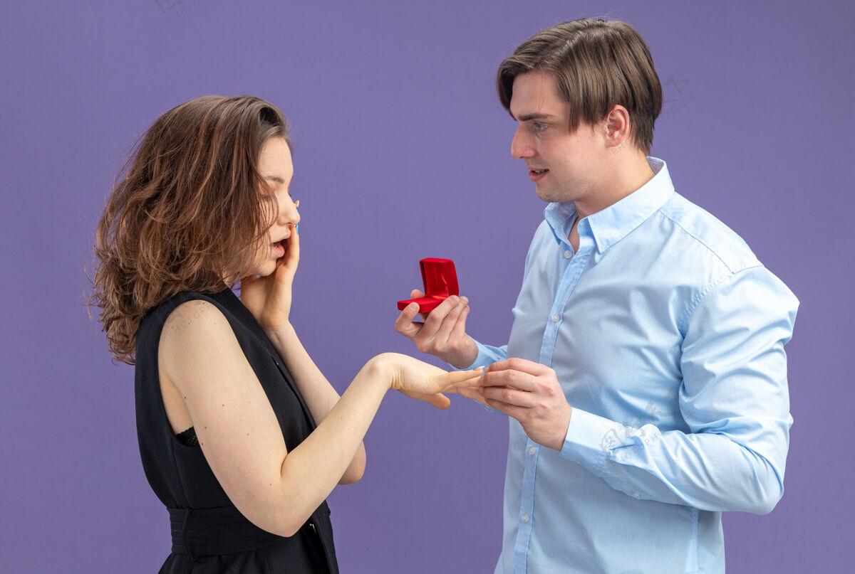 白天在情人节这天 一对年轻漂亮的情侣站在蓝色的背景下 幸福的男人拿着红盒子里的订婚戒指向他可爱而惊讶的女友求婚爱情情侣制造
