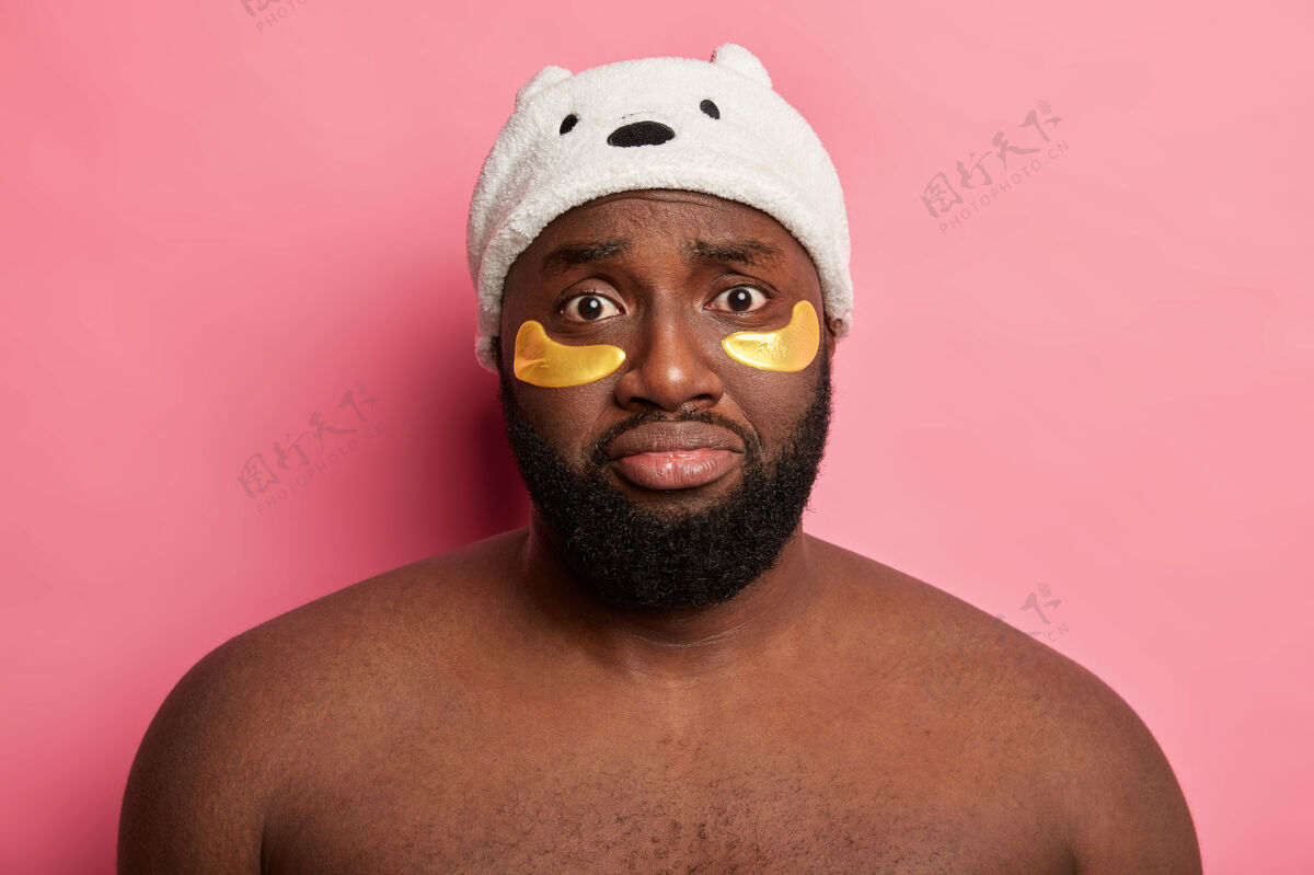 站立非洲男人伤心地看着镜头 不高兴脸上有很多皱纹 涂上金色的补丁 在家里有美容程序 隔离在粉红色的墙上水疗男性不高兴