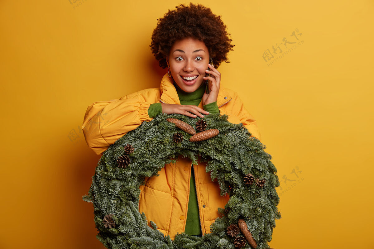 手机美丽的黑皮肤女士手持手工制作的圣诞云杉花环 有着幸福的表情 穿着黄色外套 打电话给朋友 邀请庆祝寒假 隔离在黄色背景之上科技小工具欢呼