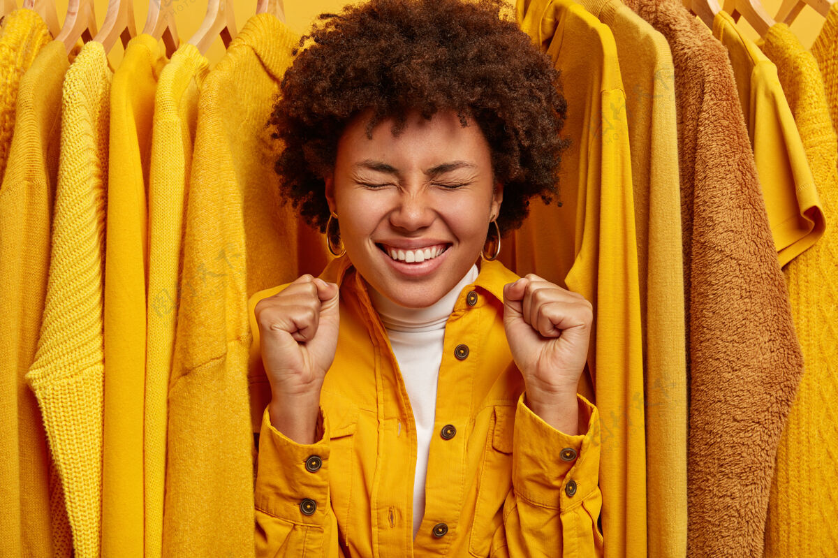 成人喜出望外的黑皮肤女人站在黄色时髦衣架旁 握紧拳头 欢欣鼓舞地购买成功优雅大笑商店