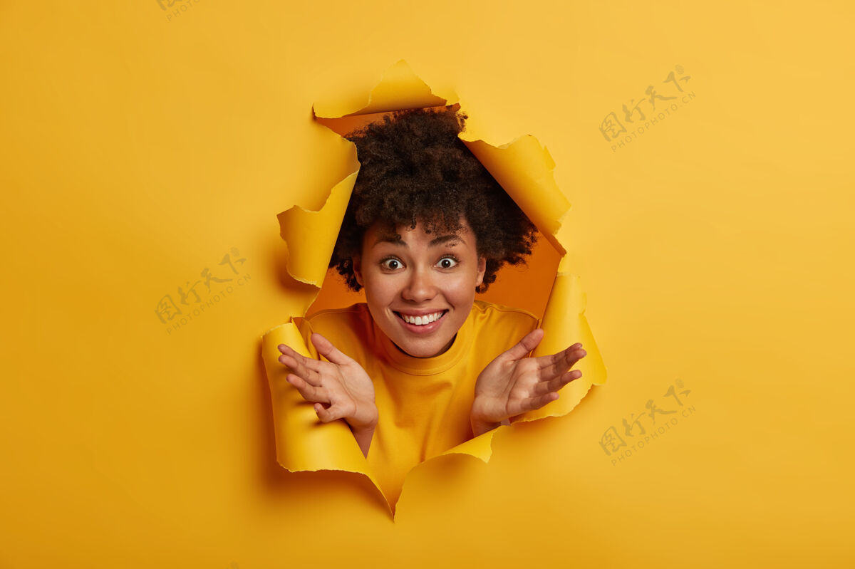 感觉快乐的非裔美国女人穿着黄色毛衣 摊开手掌 露出洁白的牙齿 在室内玩得很开心 透过撕破的纸洞看背景快乐洞情绪