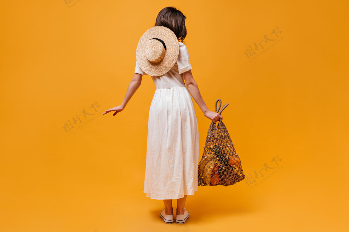 脸身着迷笛裙 头戴草帽 背着橘色背景的水果串袋的女孩的全长照片卷发长波浪发