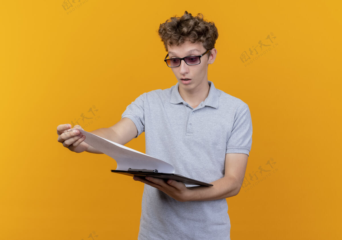 橙色一个戴着黑眼镜 穿着灰色马球衫的年轻人拿着一个空白页的剪贴板 惊讶地看着那个橘子灰色空白衬衫