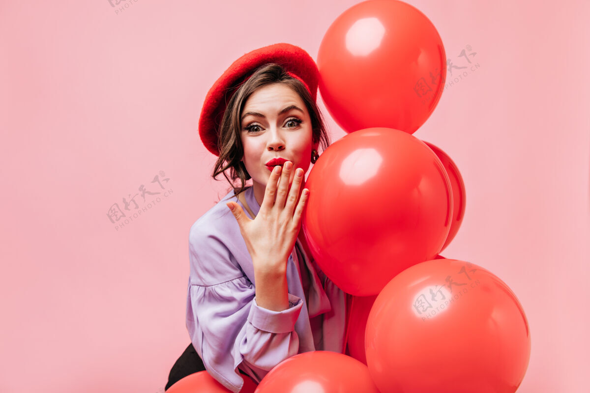 连衣裙穿着淡紫色上衣和红色贝雷帽的可爱女人吹着吻 手里拿着粉色背景上的气球漂亮微笑女孩