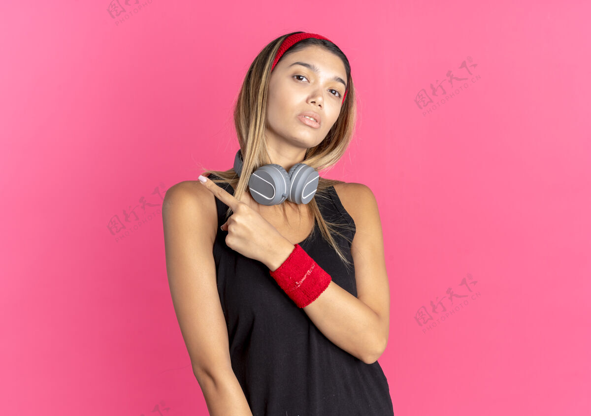 指点身穿黑色运动服 头戴红色头带 头戴耳机的年轻健身女孩看起来自信满满 手指指向粉色上方健身自信红色