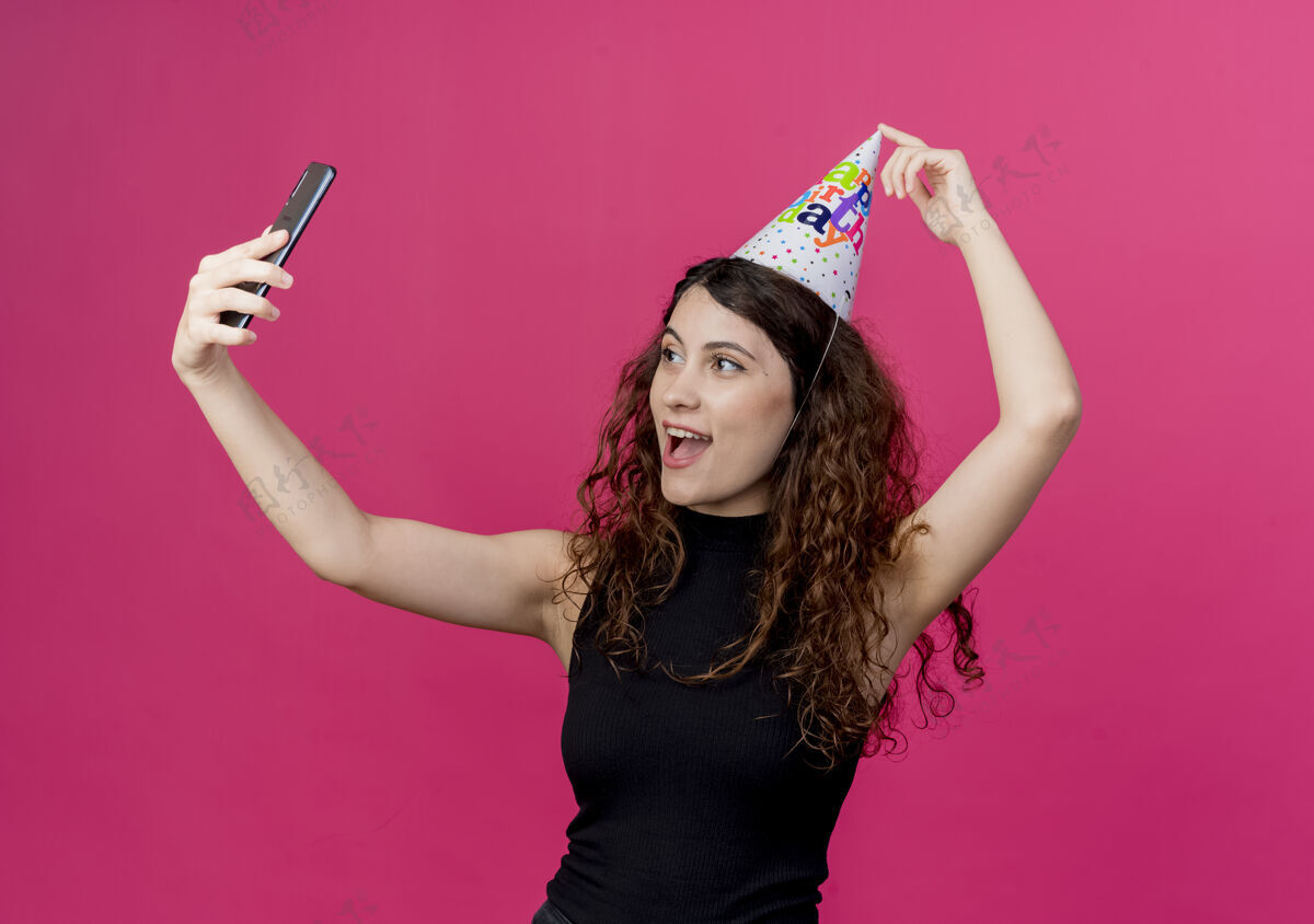 粉色年轻漂亮的女士 卷发 戴着节日帽 自拍 笑容可掬的生日派对概念 粉色拿着女人生日