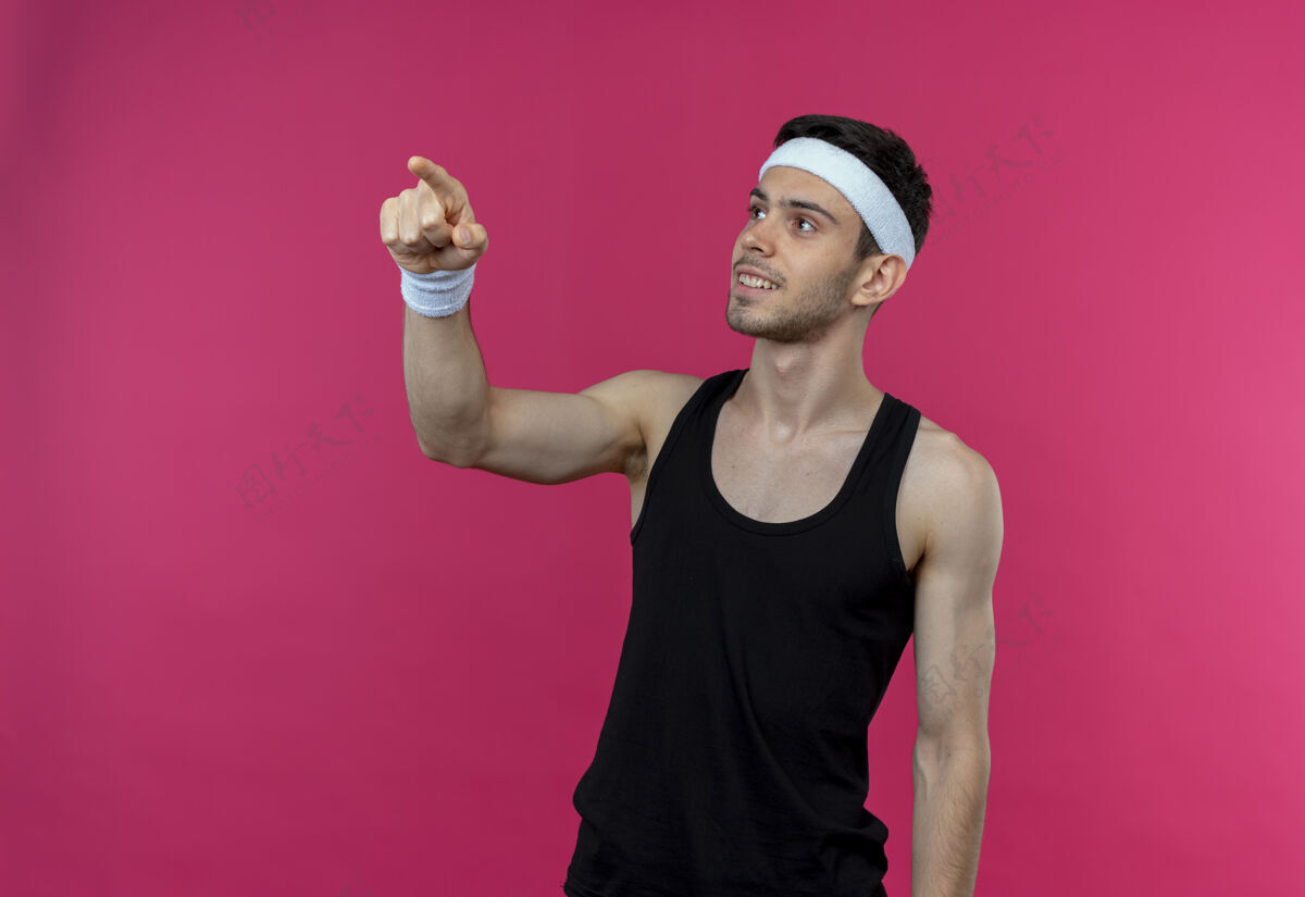 目录戴着头巾的年轻运动型男子一边看一边用食指指着粉色墙上站着的微笑的东西年轻人头带手指