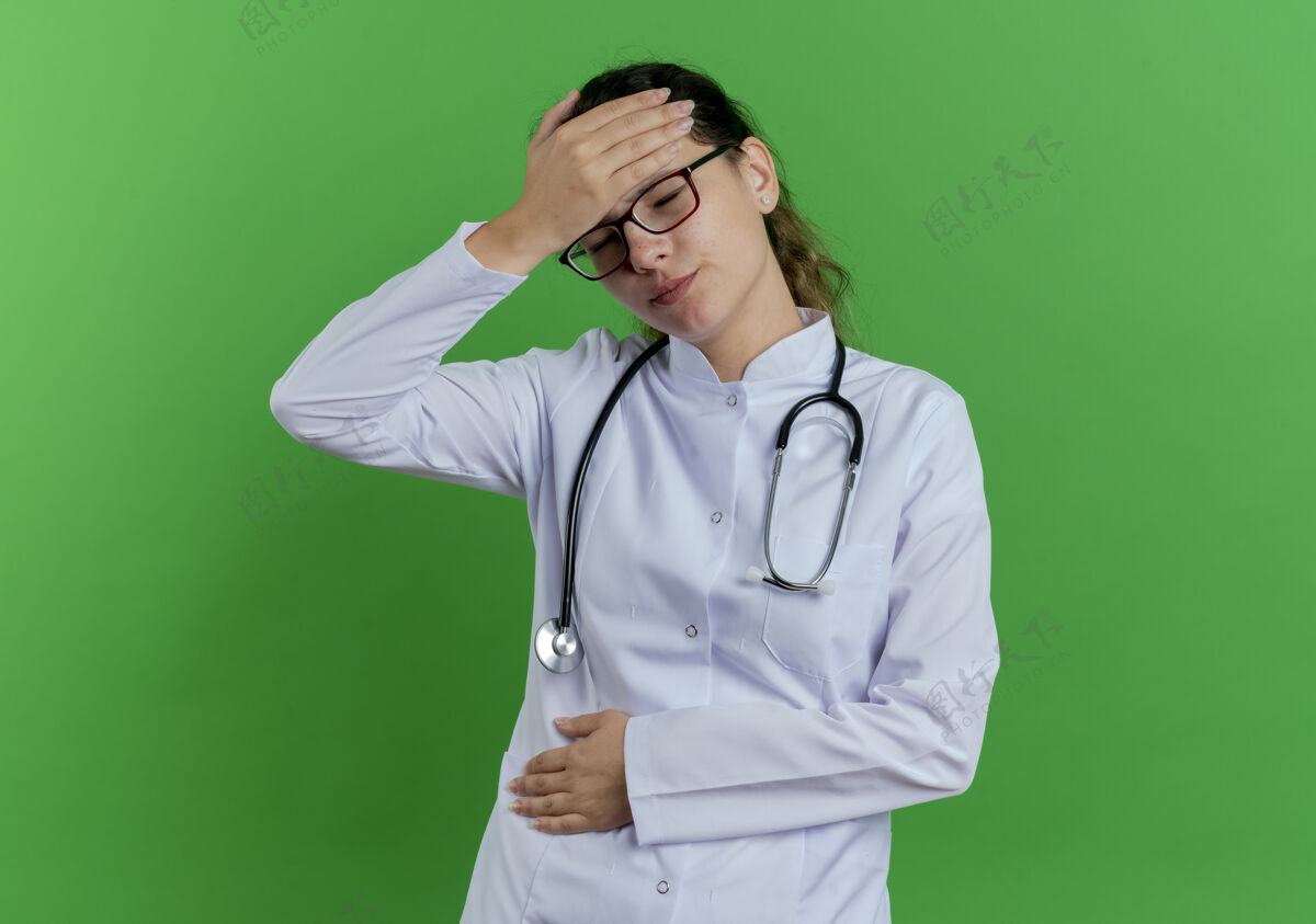 医学疼痛的年轻女医生穿着医用长袍 戴着听诊器 戴着眼镜 闭着眼睛抱着头和肚子绿色闭嘴医生