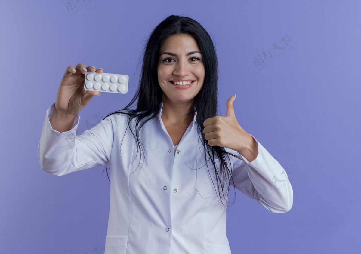 医生面带微笑的年轻女医生穿着医用长袍 展示着一包药片 抬起大拇指穿女性向上