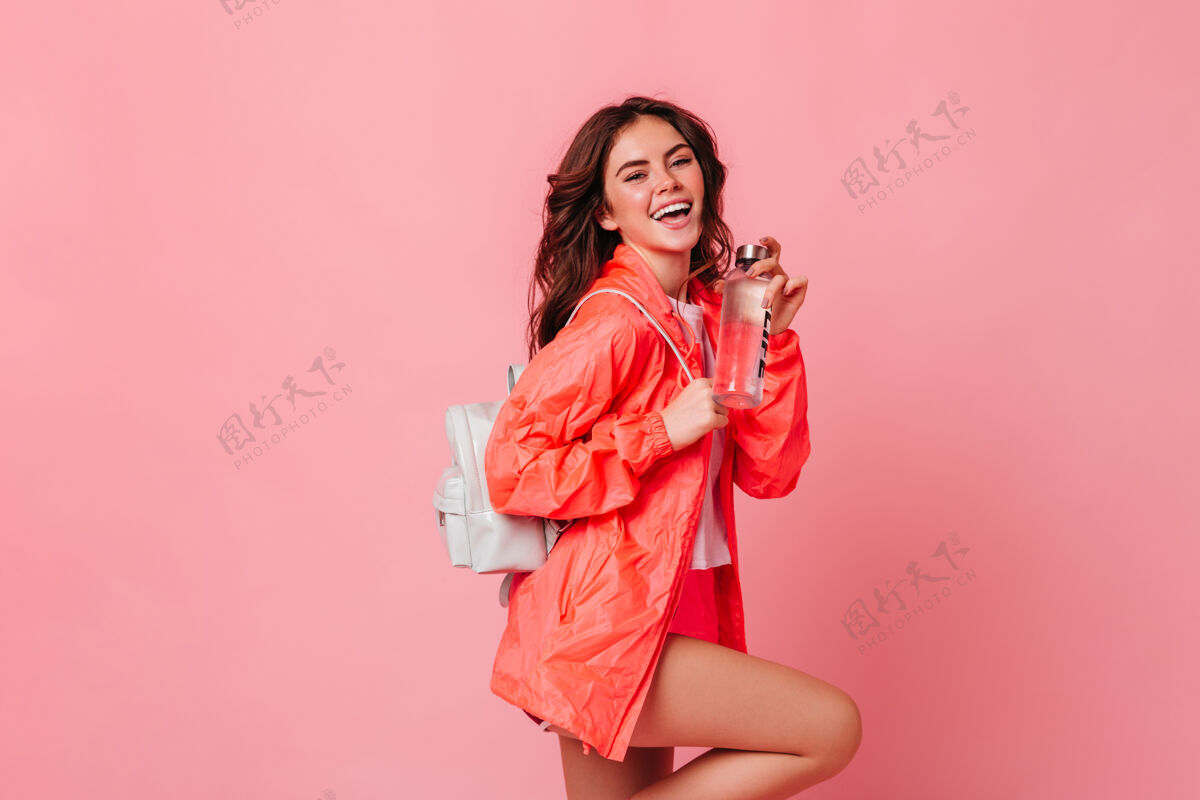 风衣穿着运动夹克 背着背包的活泼苗条的女人笑着把一瓶水放在粉红色的墙上嘴唇运动风格卷发