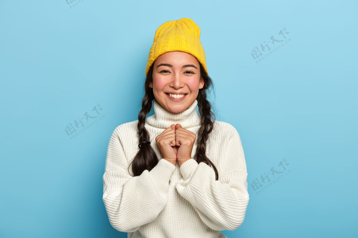 快乐快乐少女的画像双手合十 对着镜头微笑 享受好消息 戴着黄色帽子和白色大号毛衣人快乐头饰
