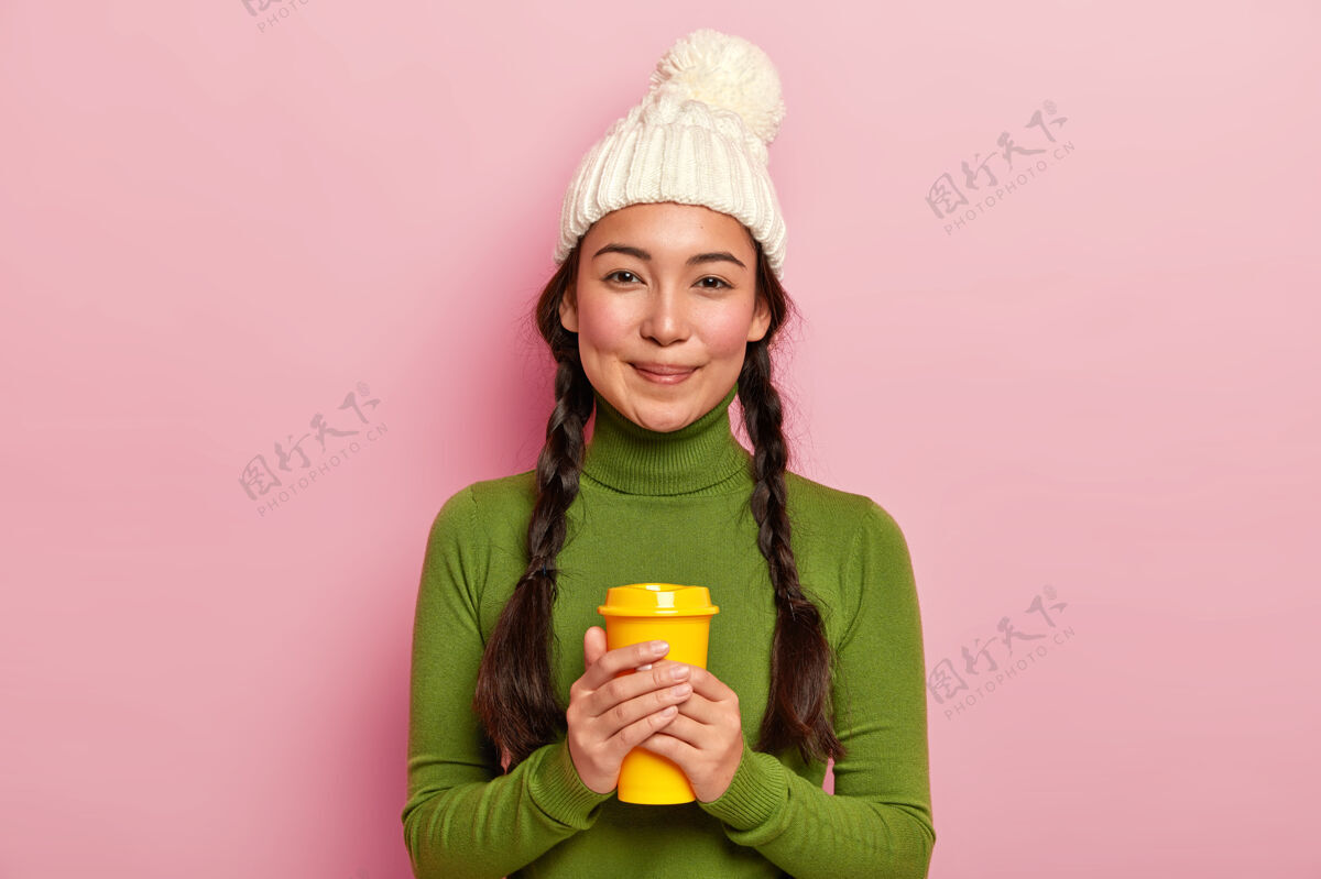 茶放松可爱的黑头发女人 留着两条辫子 拿着外卖咖啡 冬天用热饮料取暖 戴着白帽子和绿色高领毛衣欢呼时尚亚洲人