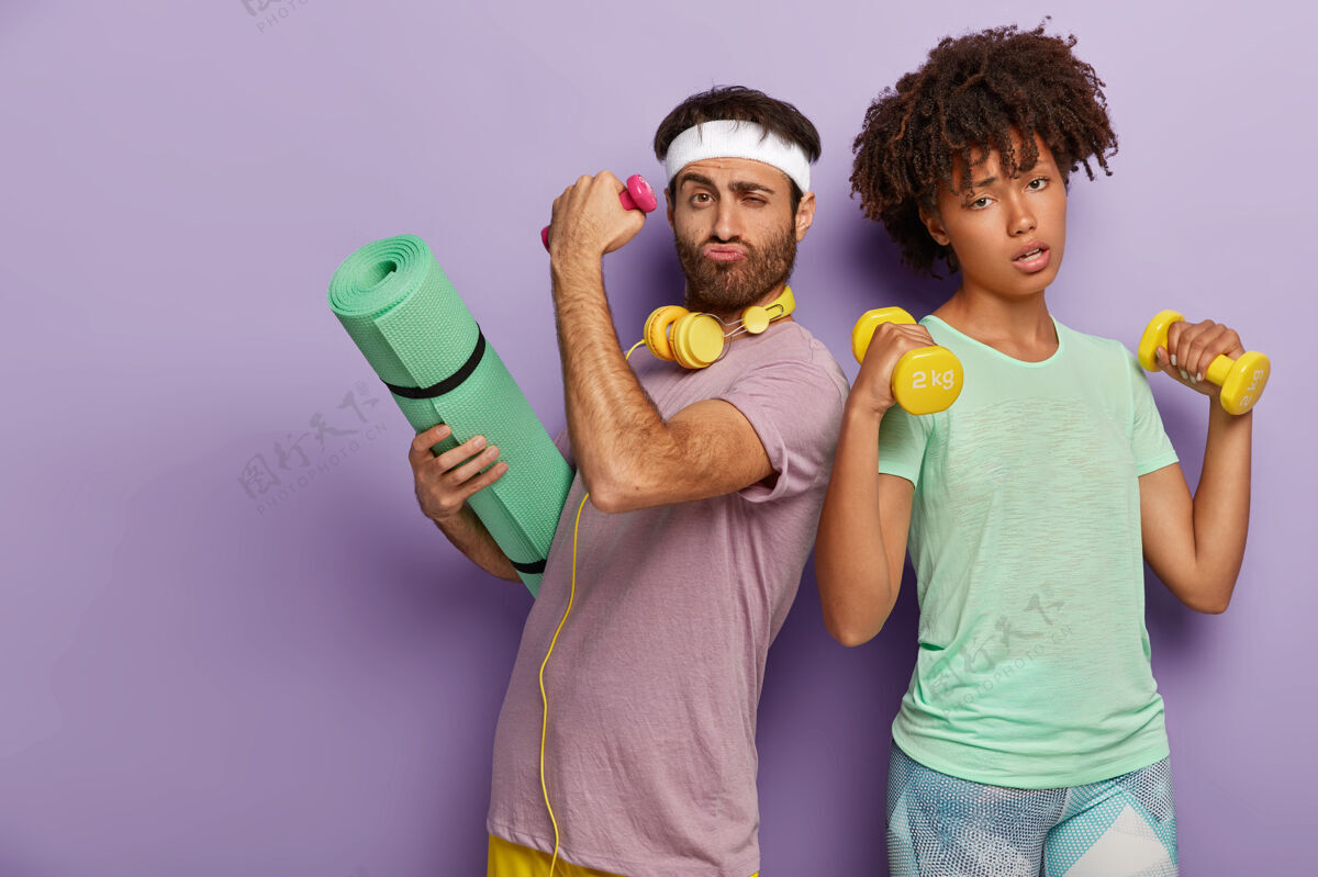 健康疲惫的女人举起两个哑铃 锻炼肱二头肌 决心不刮胡子的男人背着卡雷马特进行体操训练设备耳机男