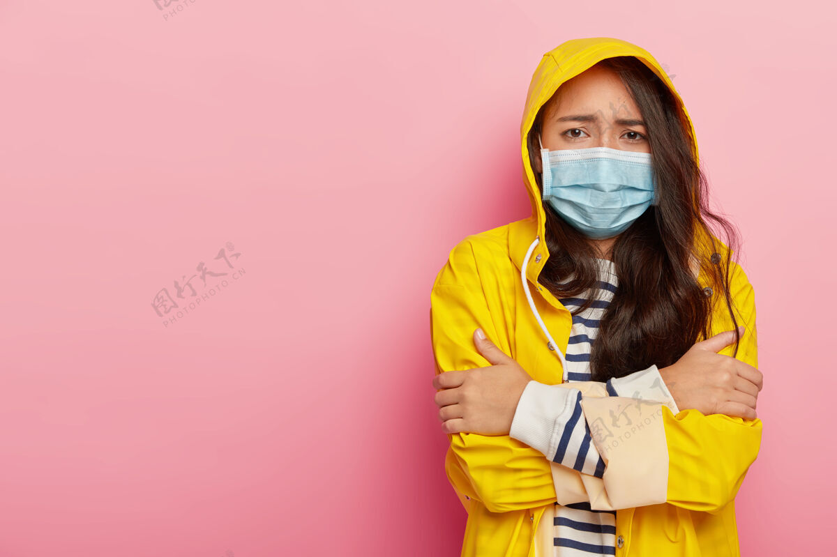 条纹心烦意乱的亚洲妇女因寒冷而发抖 病毒通过空气中的飞沫传播 戴着防护性的医用口罩 黄色雨衣和风帽季节心烦衣服