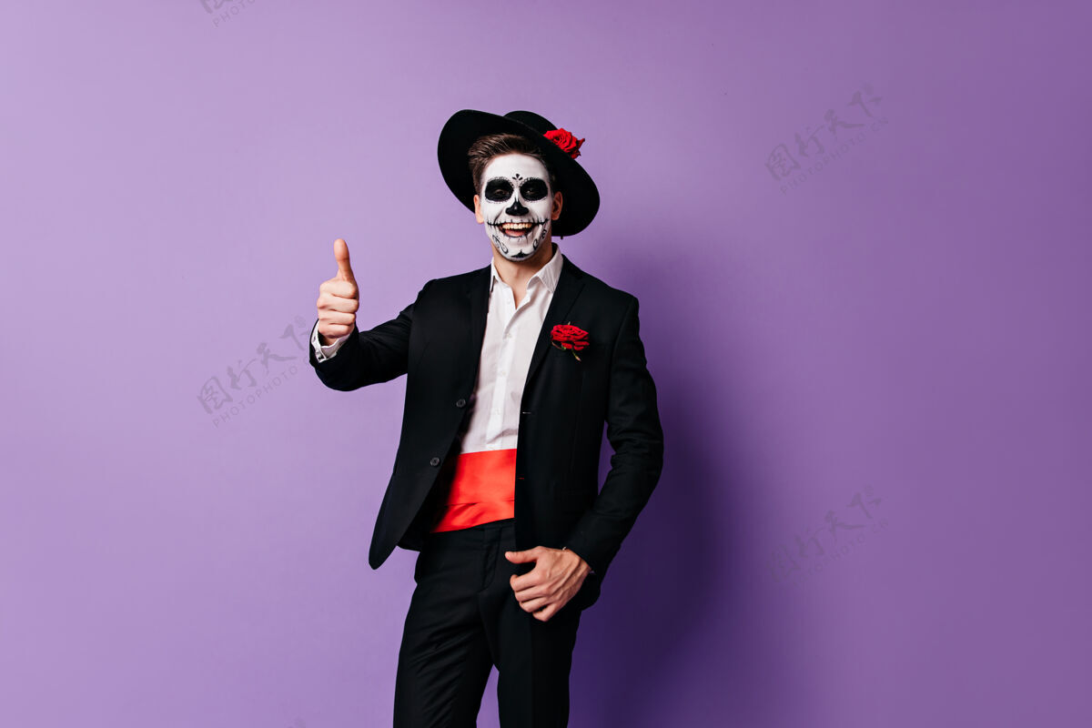 传统一个穿着墨西哥衣服戴着面具的快乐男人笑着在紫色的背景上竖起大拇指红色玫瑰幽灵
