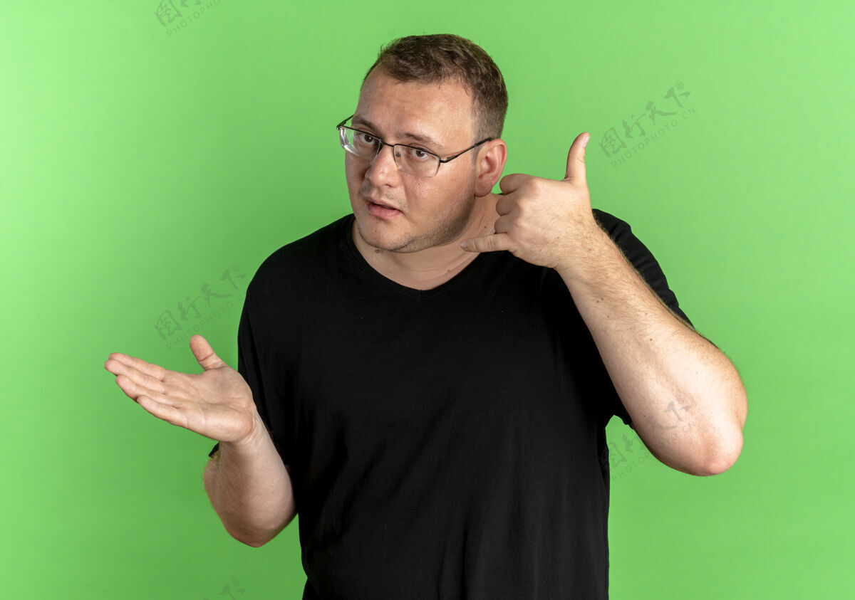 制造戴眼镜的超重男子穿着黑色t恤 看起来很困惑 在绿色上面做着叫我的手势绿色表情T恤