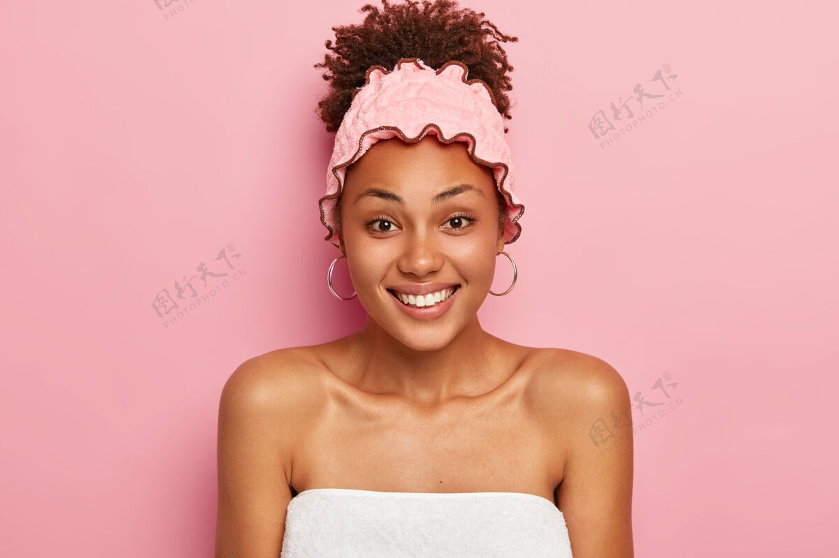 欢呼快乐华丽的非洲裔女士微笑着 裹着毛巾 戴着柔软的浴帽 享受着洗澡 隔离在粉红色的墙上健康头部情感