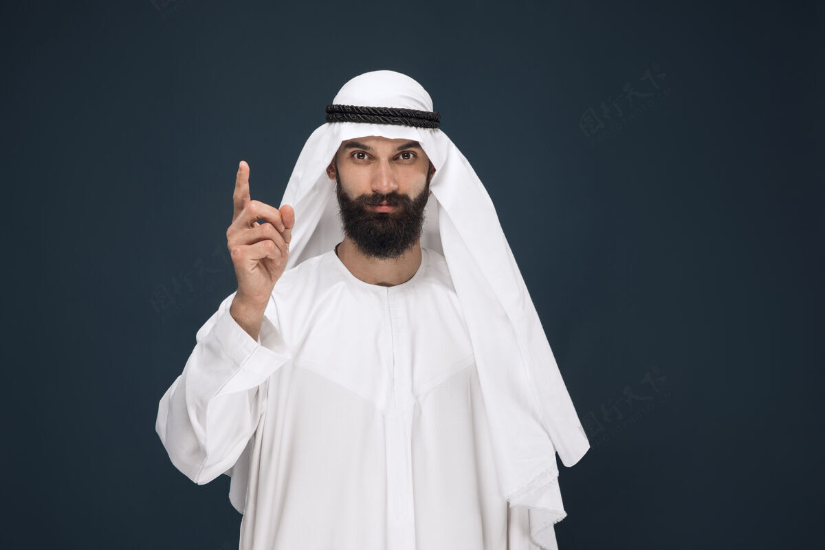 胡须深蓝色墙上的半身阿拉伯沙特商人肖像年轻的男模微笑着指着商业 金融 面部表情 人类情感的概念面部衣服阿联酋