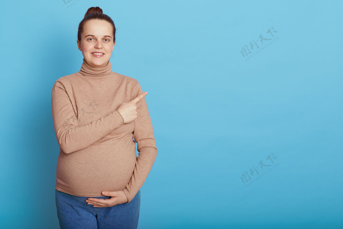 腹部欧洲年轻漂亮的孕妇 一只手摸着肚子 食指指向一边 孤零零地站在蓝色的墙上 抱着发髻的准妈妈成人壁板欢呼