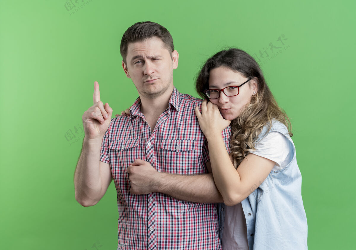 女人一对穿着休闲服的年轻夫妇站在一起 自信的男人在绿墙上展示指数斗鸡男人立场年轻人