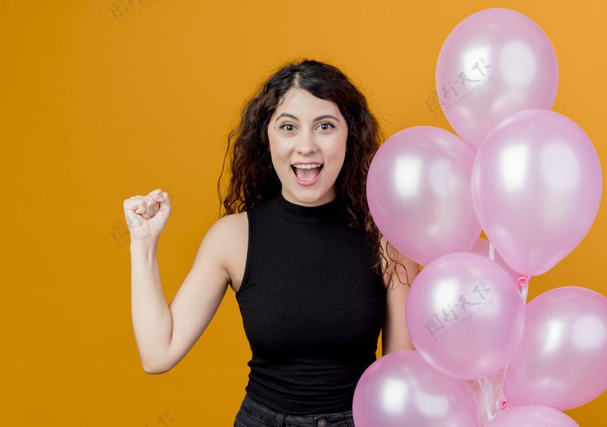 举行年轻漂亮的卷发女子手持一束气球握紧拳头站在橙色的墙上 快乐而兴奋的生日派对概念气球年轻站着
