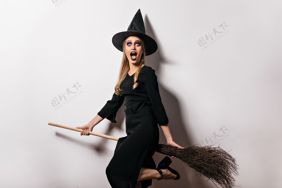 美丽震惊女巫与黑色化妆摆在万圣节迷人的金发女郎在魔法帽子拿着扫帚享受年轻女人