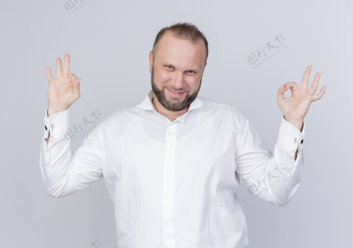站着留着胡子的男人穿着白衬衫 开心而积极地微笑着 站在白墙上做着“ok”的手势男人积极的穿
