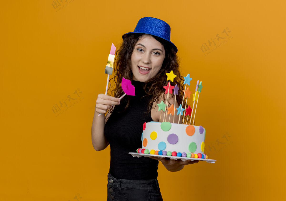 站着一位年轻漂亮的卷发女士 戴着节日礼帽 手里拿着生日蛋糕 站在橘色的墙上 开心而积极地微笑着女人蛋糕年轻