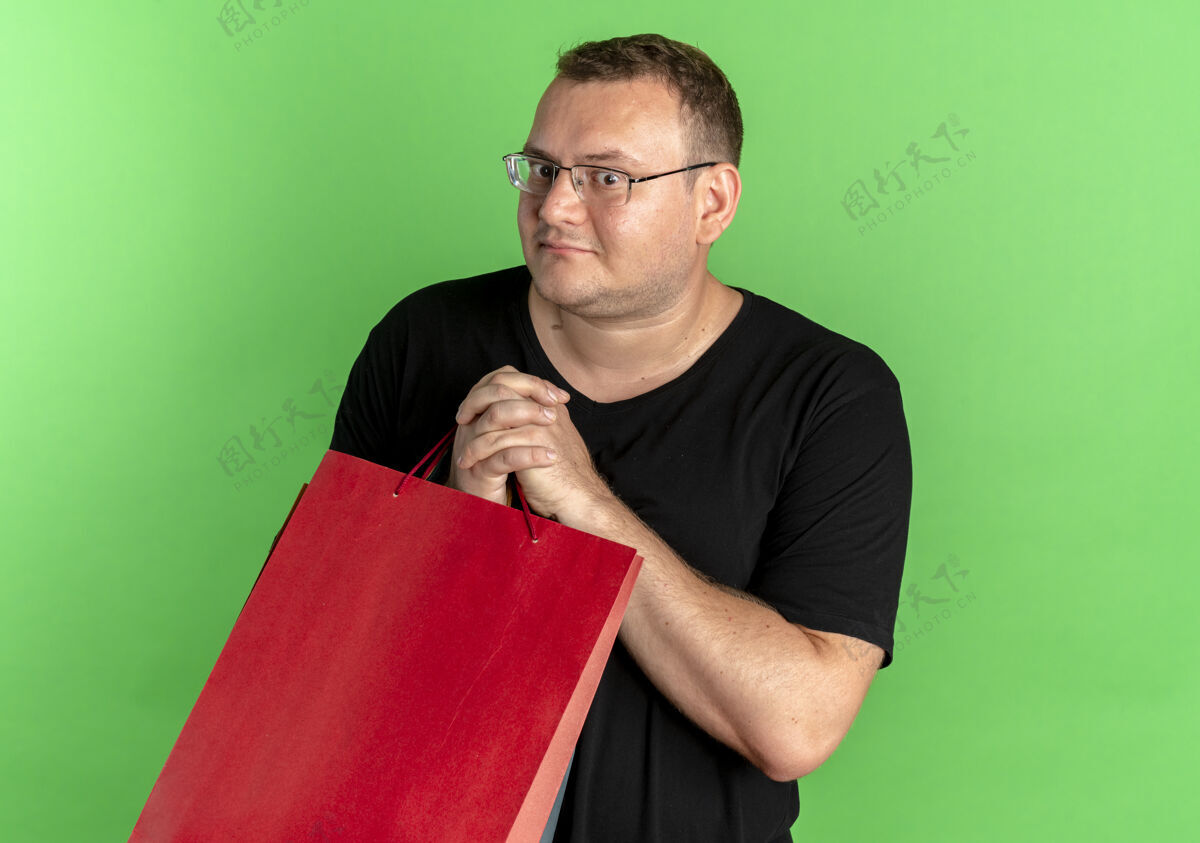 男人一个戴眼镜 身穿黑色t恤 手拿纸袋的超重男子站在绿色的墙上 看上去很困惑超重站穿