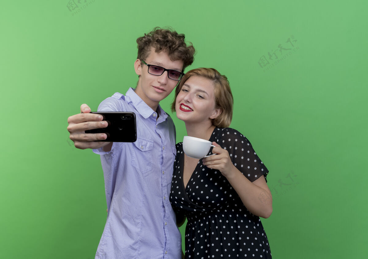 微笑年轻漂亮的情侣站在一起用手机自拍微笑着越过绿墙用着自拍年轻