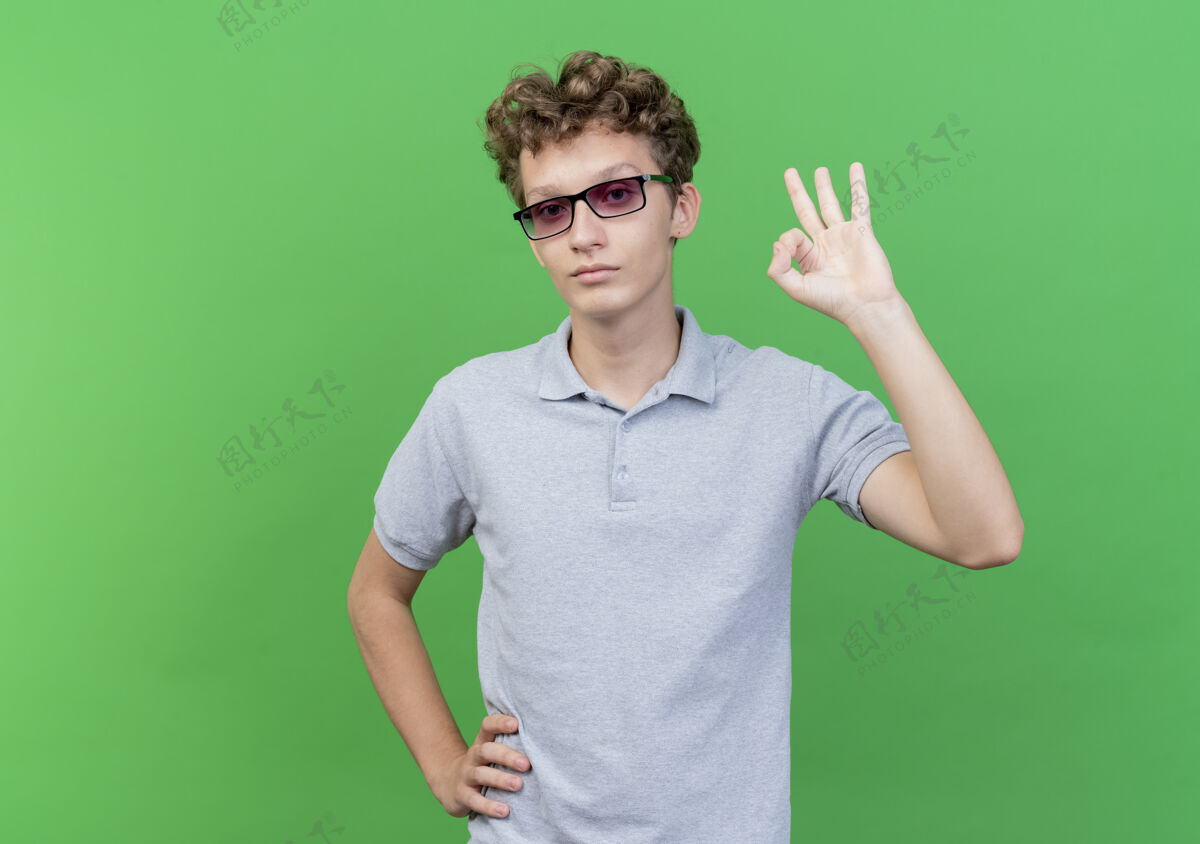 穿戴着黑眼镜 身穿灰色马球衫的年轻人站在绿色的墙上 高兴而积极地展示着ok标志看站马球