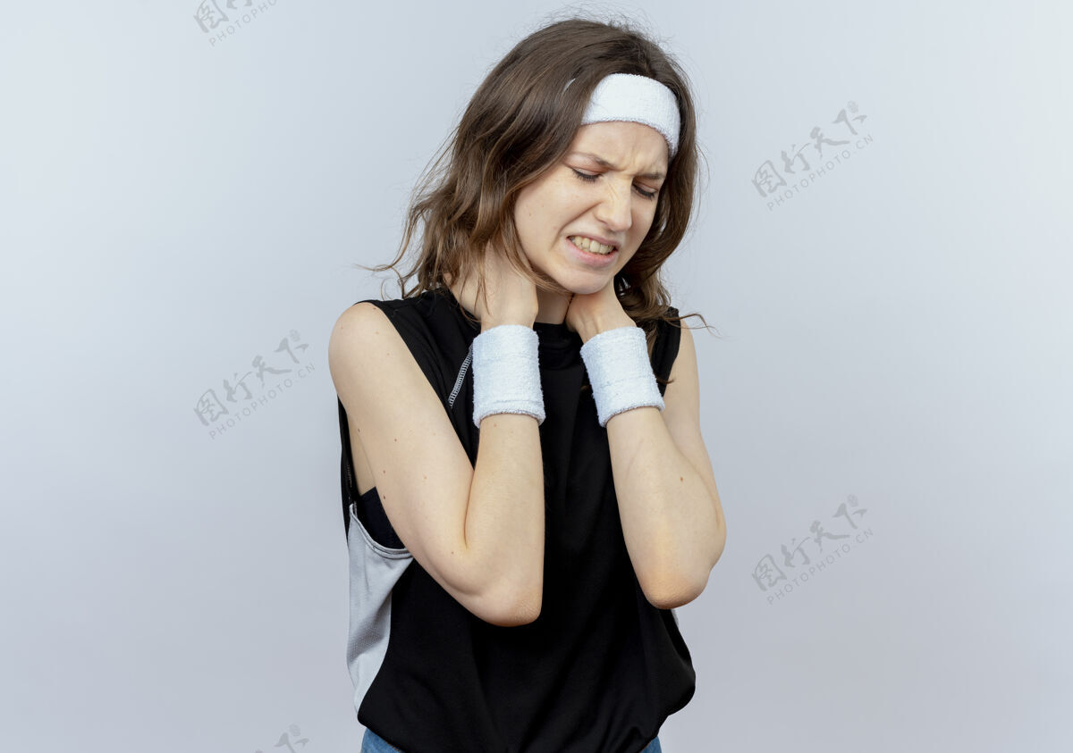 触摸穿着黑色运动服的年轻健身女孩戴着头带 看起来不舒服 站在白墙上摸着脖子疼头带疼痛健康