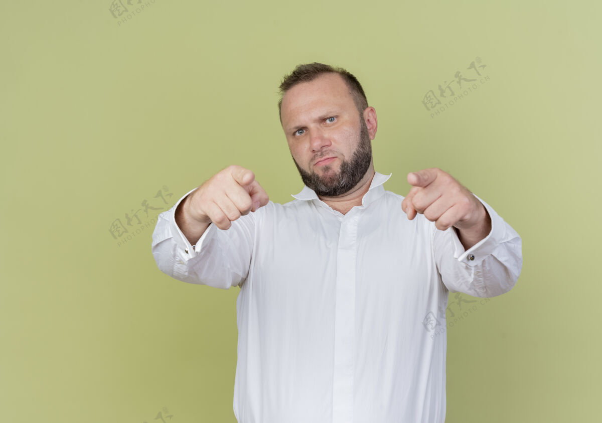 胡子一个留着胡子的男人穿着白衬衫 用食指指着 站在轻质的墙上 看起来很不高兴光人指