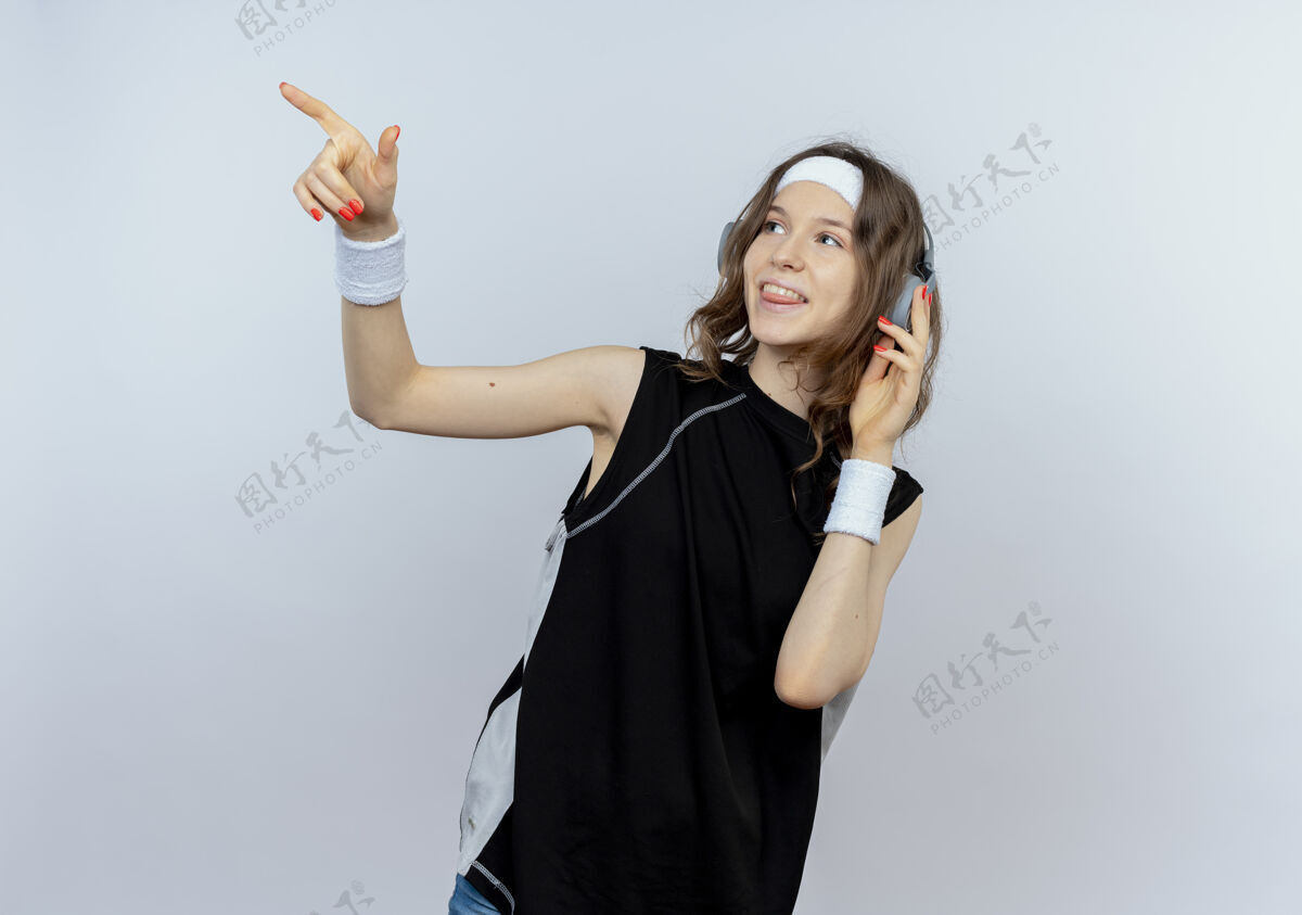 手指身穿黑色运动服的年轻健身女孩戴着头带和耳机 面带微笑 手指着站在白墙上的东西放一边女孩微笑