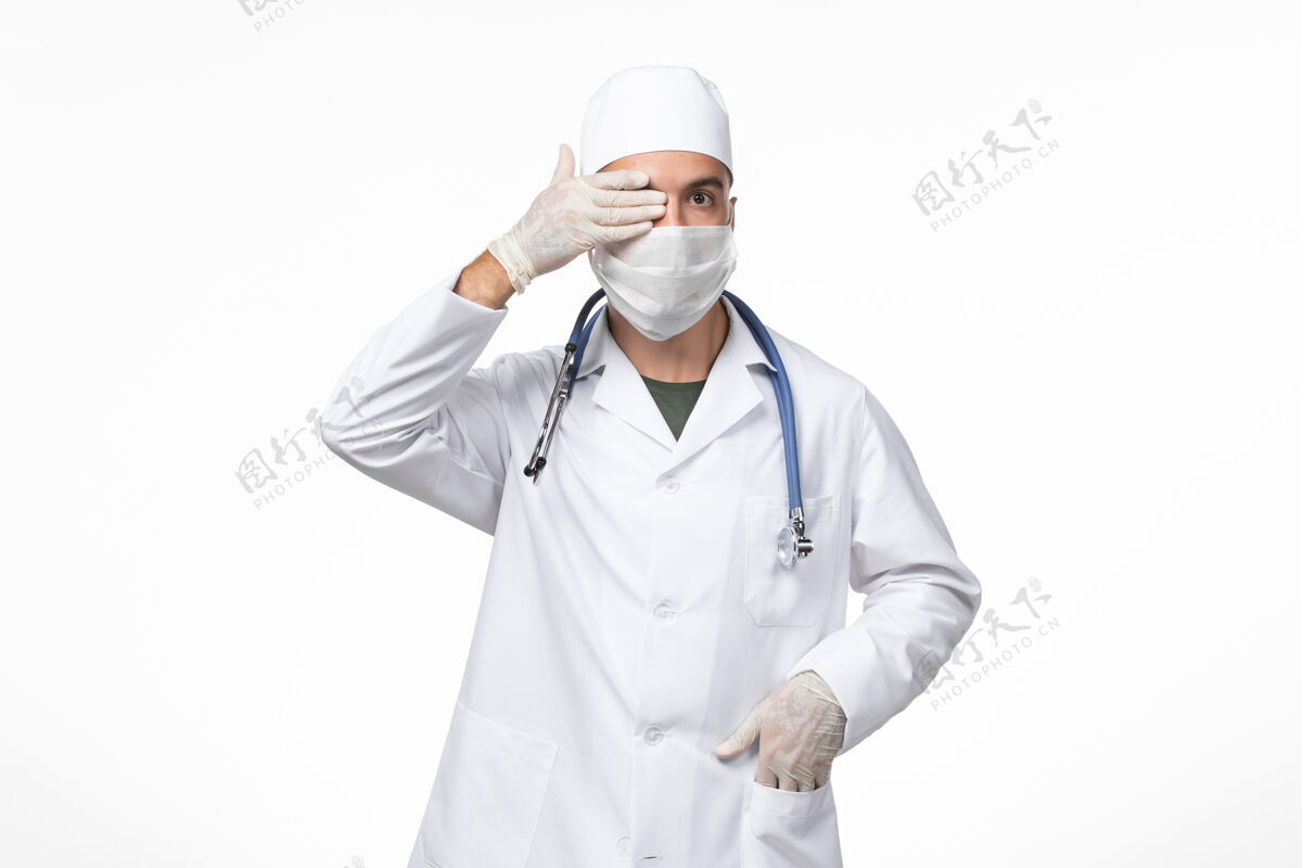 男性正面图：男医生穿着医疗服 戴着防冠状病毒口罩 浅白色墙壁上有蓝色听诊器 冠状病毒-疾病大流行病毒西装病毒男人