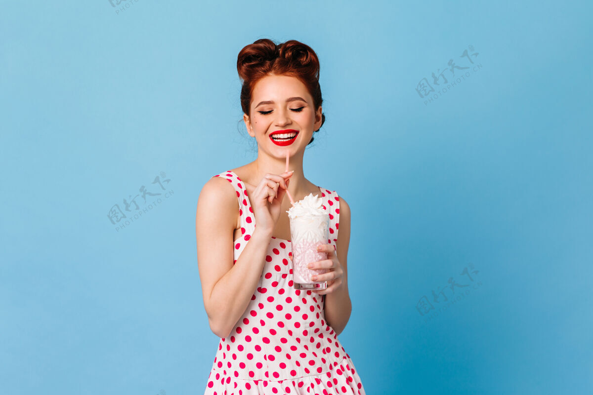 波尔卡多迷人的姜汁女孩喝着奶昔摄影棚拍摄的穿着圆点裙的快乐女士在蓝色空间享受饮料发型黑发魅力