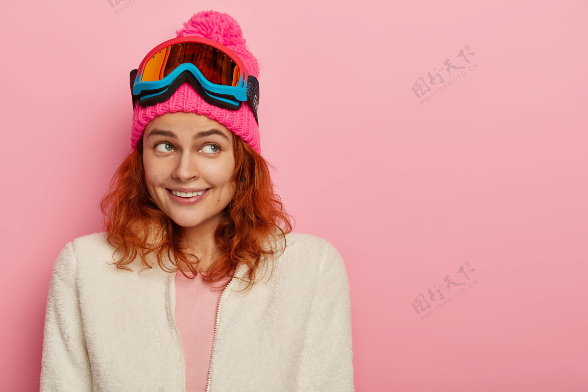 微笑快乐的滑雪者女孩微笑着温柔 专注在一边 戴着粉色的蓬蓬冬帽 拉链上套着白色软毛衣 头上戴着滑雪眼镜 对着玫瑰色的墙壁摆姿势姜女人室内