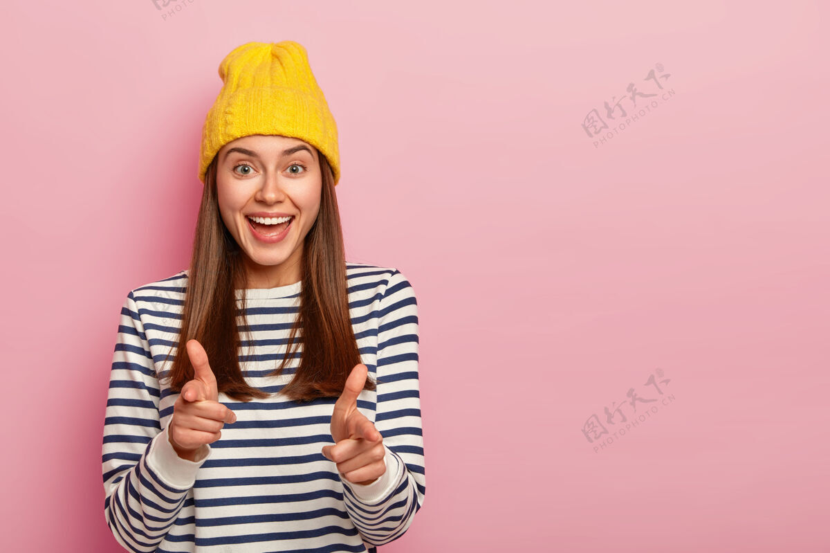 复制空间迷人的年轻女性穆德尔的照片做手指枪手势 戴黄色帽子和条纹套头衫 表达选择 选择你到她的团队 孤立在粉红色背景上显示套头衫微笑