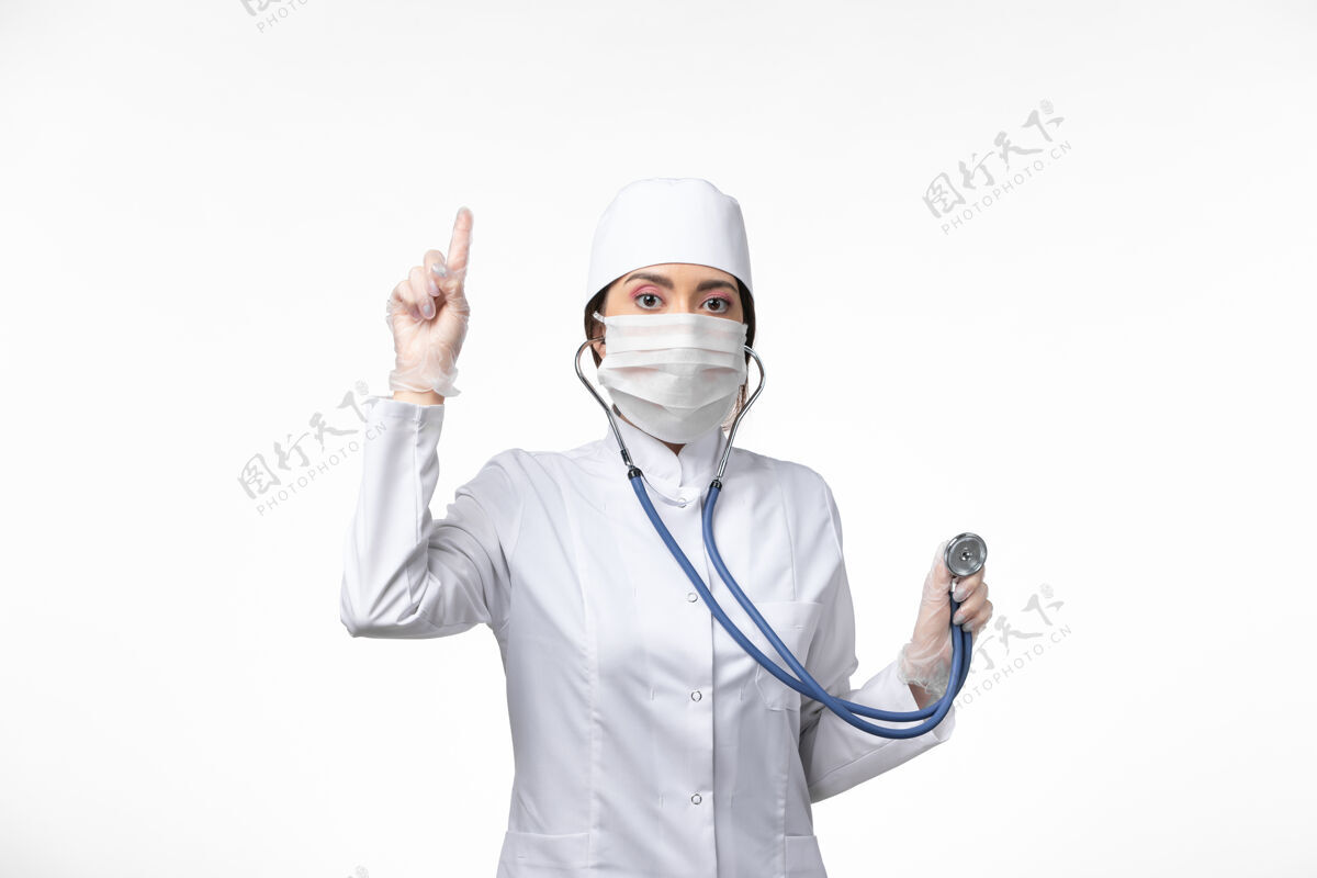 套装正面图：女医生穿着白色无菌医疗服 戴口罩 因冠状病毒-使用听诊器对白色桌面疾病病毒冠状病毒-大流行性疾病口罩使用医生