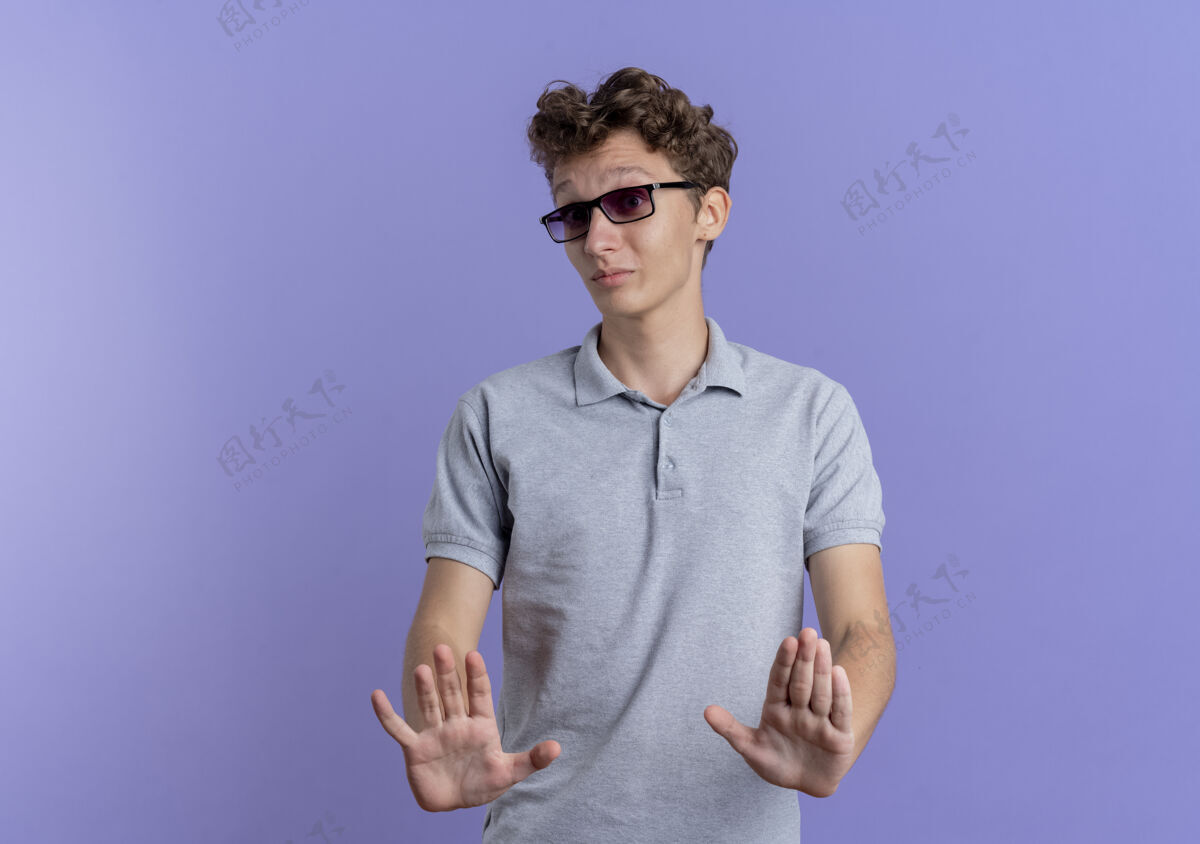 男人一个戴着黑眼镜 穿着灰色马球衫的年轻人站在蓝色的墙上 手举着手示意不要靠近穿做抱着